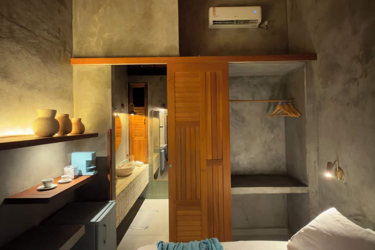 A compact bedroom with a bed, a wooden wardrobe, a small desk with a mirror, and a shelf with decorative items.