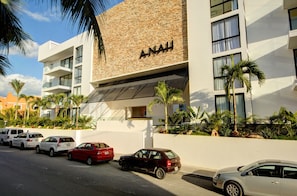Et moderne hotel med facade i mursten, flere balkoner og en parkeringsplads med flere biler.
