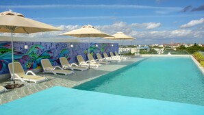 A poolside area with lounge chairs and umbrellas, a colorful mural, and a view of buildings in the distance. - Anah Suites - Las Flores Properties (Playa del Carmen)