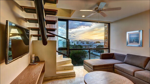 A modern living room with a staircase, a large window with a city view, a ceiling fan, and a framed picture on the wall. - Anah Suites - Las Flores Properties (Playa del Carmen)