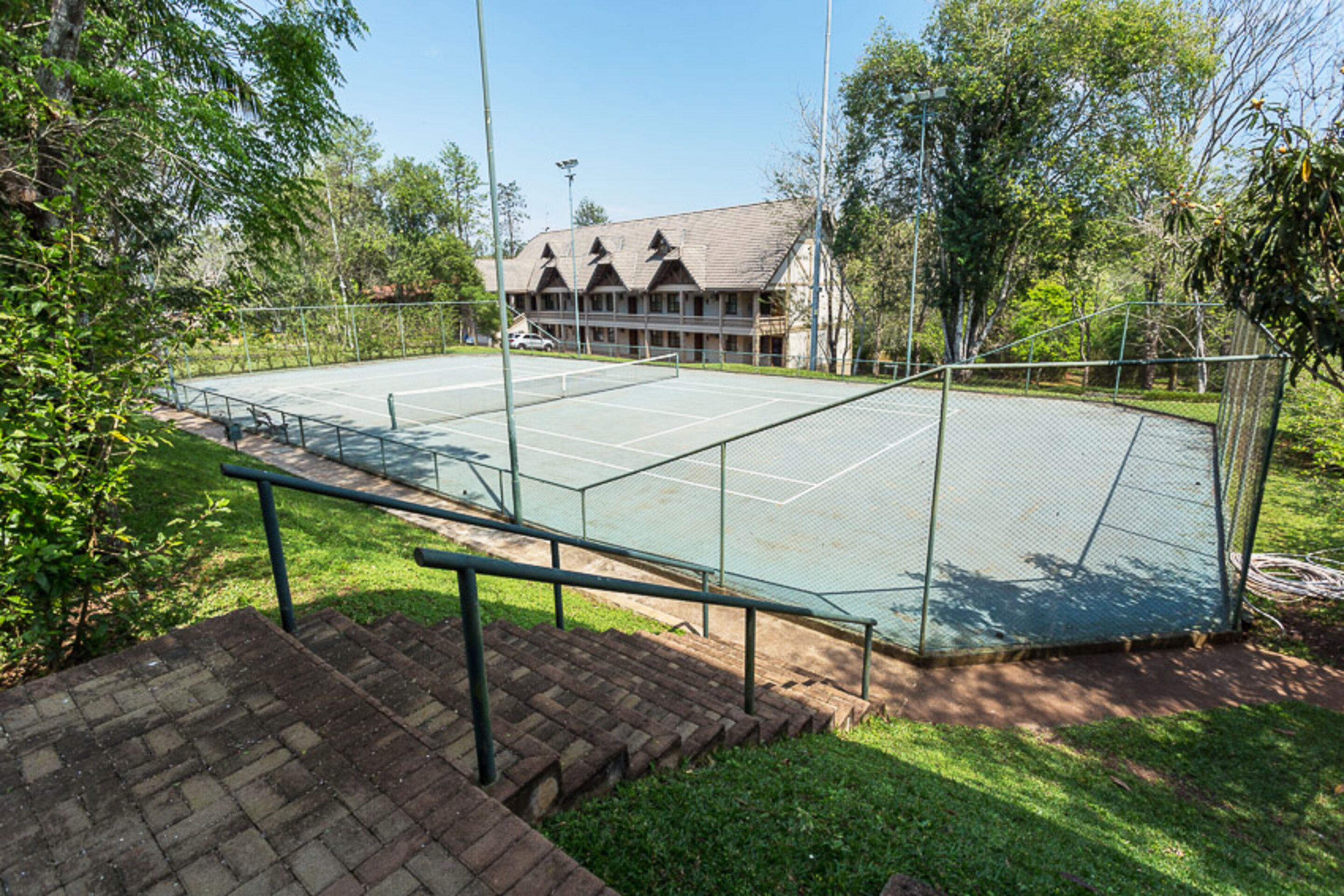 An outdoor tennis court with a net, surrounded by a fence, and a building with a sloped roof in the background.