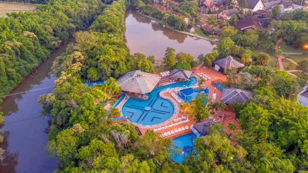An aerial view of a resort with a central swimming pool, surrounded by lush greenery and a river.