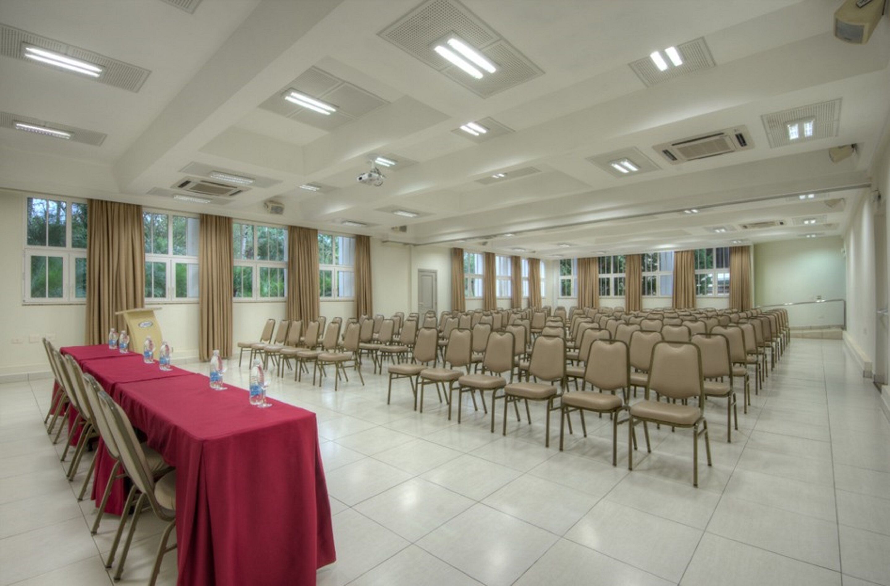 Uma grande sala de conferências com fileiras de cadeiras bege, uma mesa comprida com toalha vermelha e janelas amplas com cortinas.