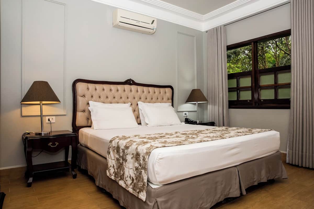 Ein ordentlich eingerichtetes Schlafzimmer mit einem großen Bett, zwei Nachttischen, einem Fenster mit Vorhängen und einer Klimaanlage an der Wand.