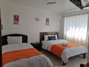A hotel room with two beds, each with white bedding and orange accents, a wooden headboard, a nightstand, and a wall-mounted light fixture. - Qori punku - Hostel (Cusco)