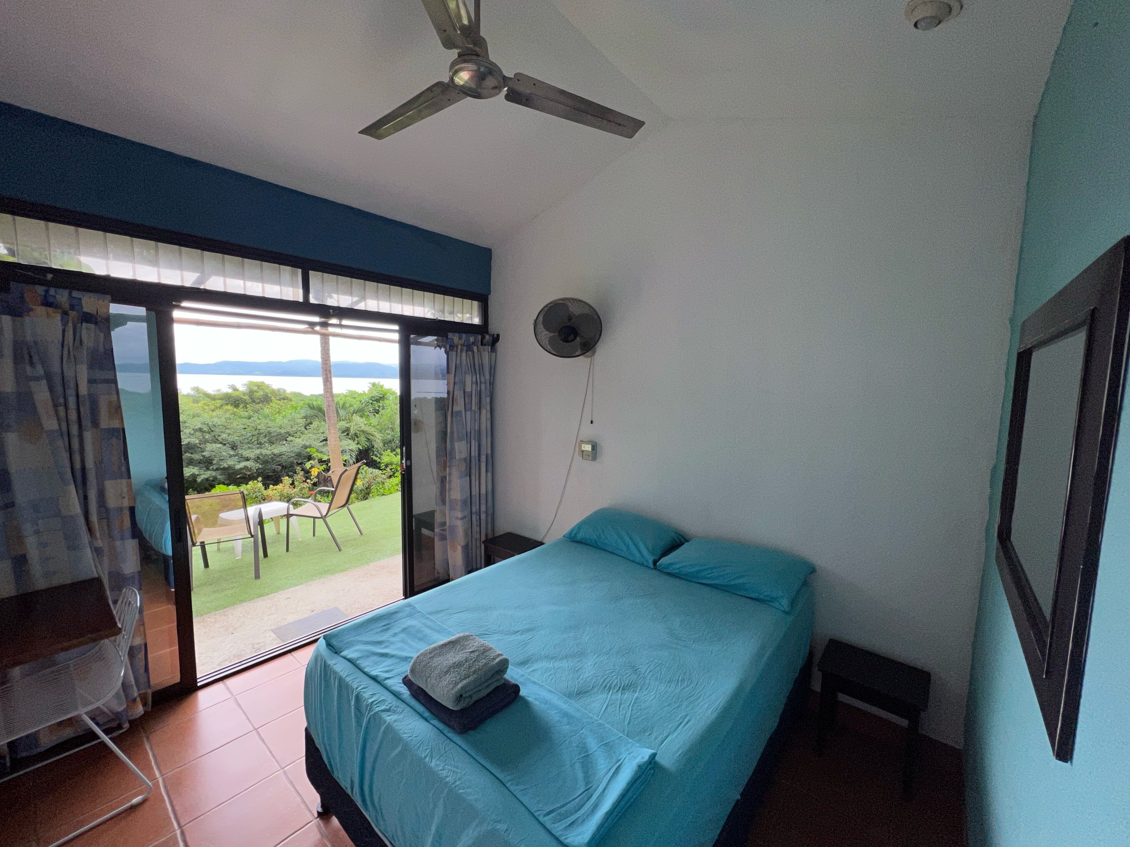 Ein Schlafzimmer mit einem Bett, einem Deckenventilator, Blick ins Freie und einer Schiebeglastür.