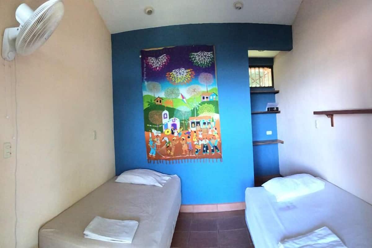 Une chambre avec deux lits, un ventilateur fixé au mur et une fresque murale aux couleurs vives.