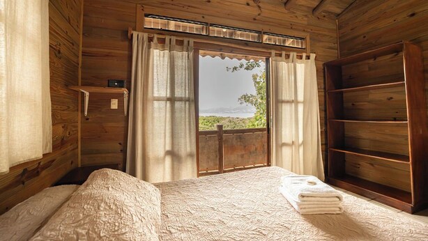 Una habitación de cabaña de madera con una cama, una ventana con cortinas y un estante de madera.