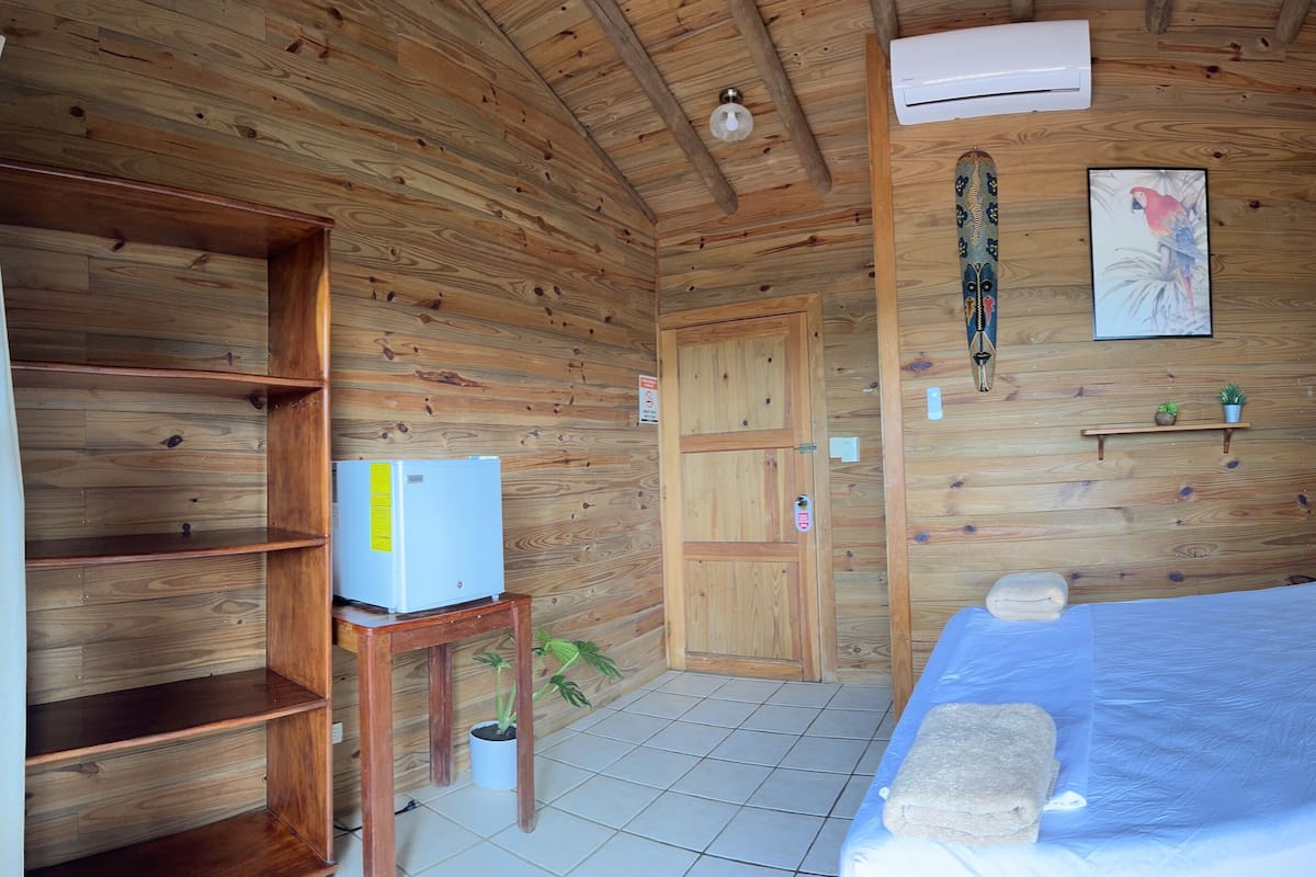 Une pièce de cabane en bois comprenant un lit, une petite table avec un réfrigérateur, une porte et une fenêtre avec des rideaux.