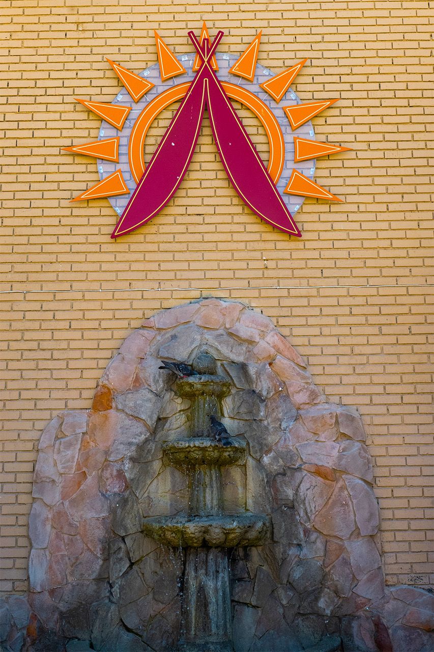 A stone fountain with a decorative arch, a sun emblem, and a pair of red ribbons on a brick wall.