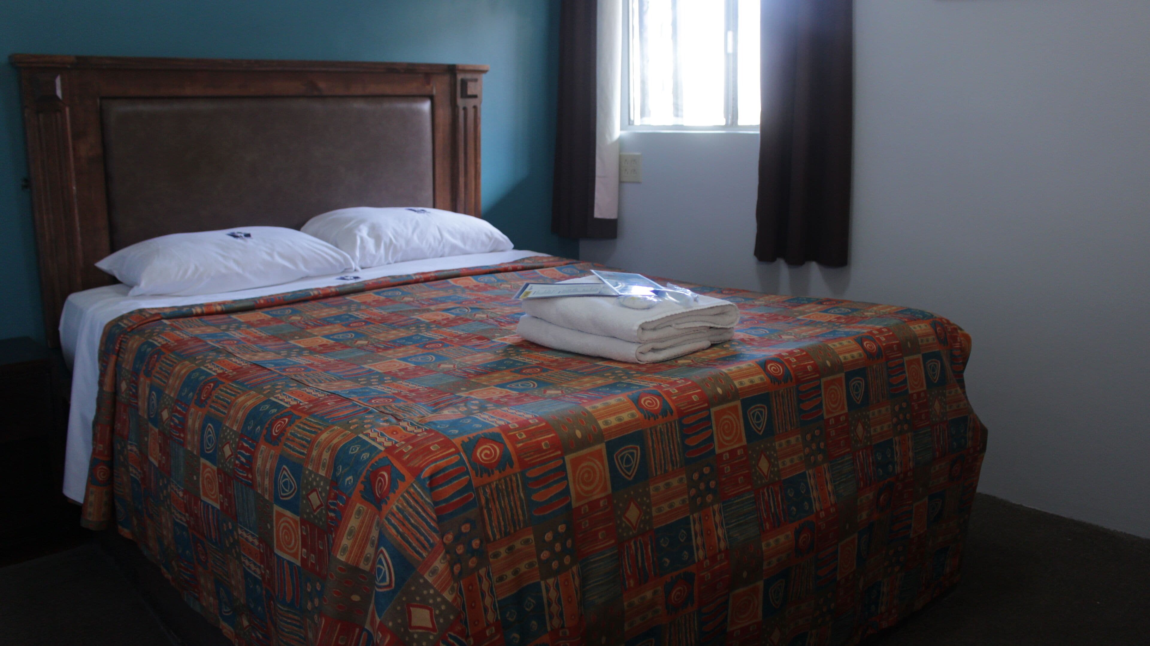 A neatly made bed with a colorful patterned bedspread and white pillows, a wooden headboard, and a window with curtains.