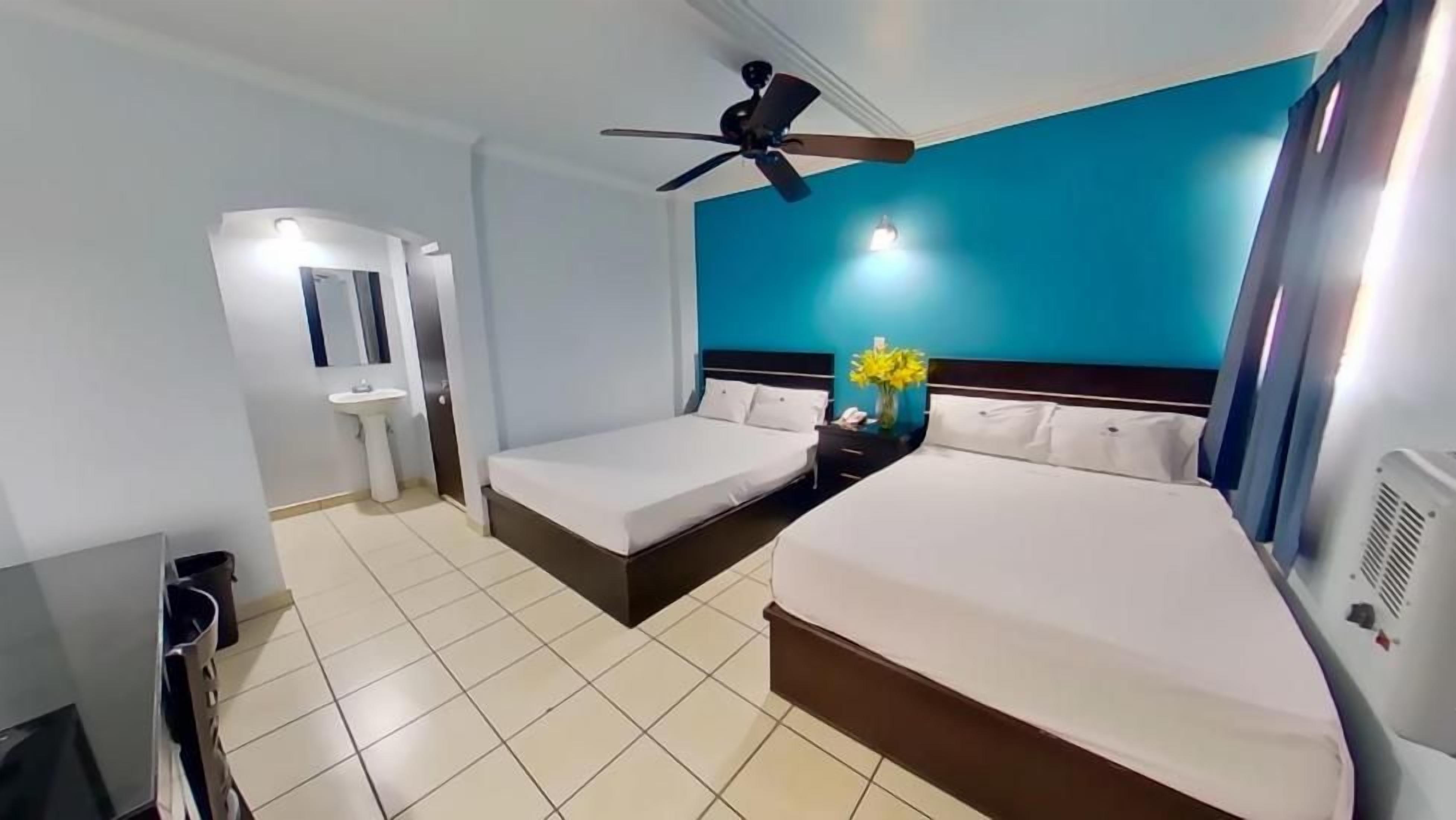 Quarto de hotel com cama, mesa de cabeceira, ventoinha no teto, TV fixada na parede e banheiro com pia e espelho.