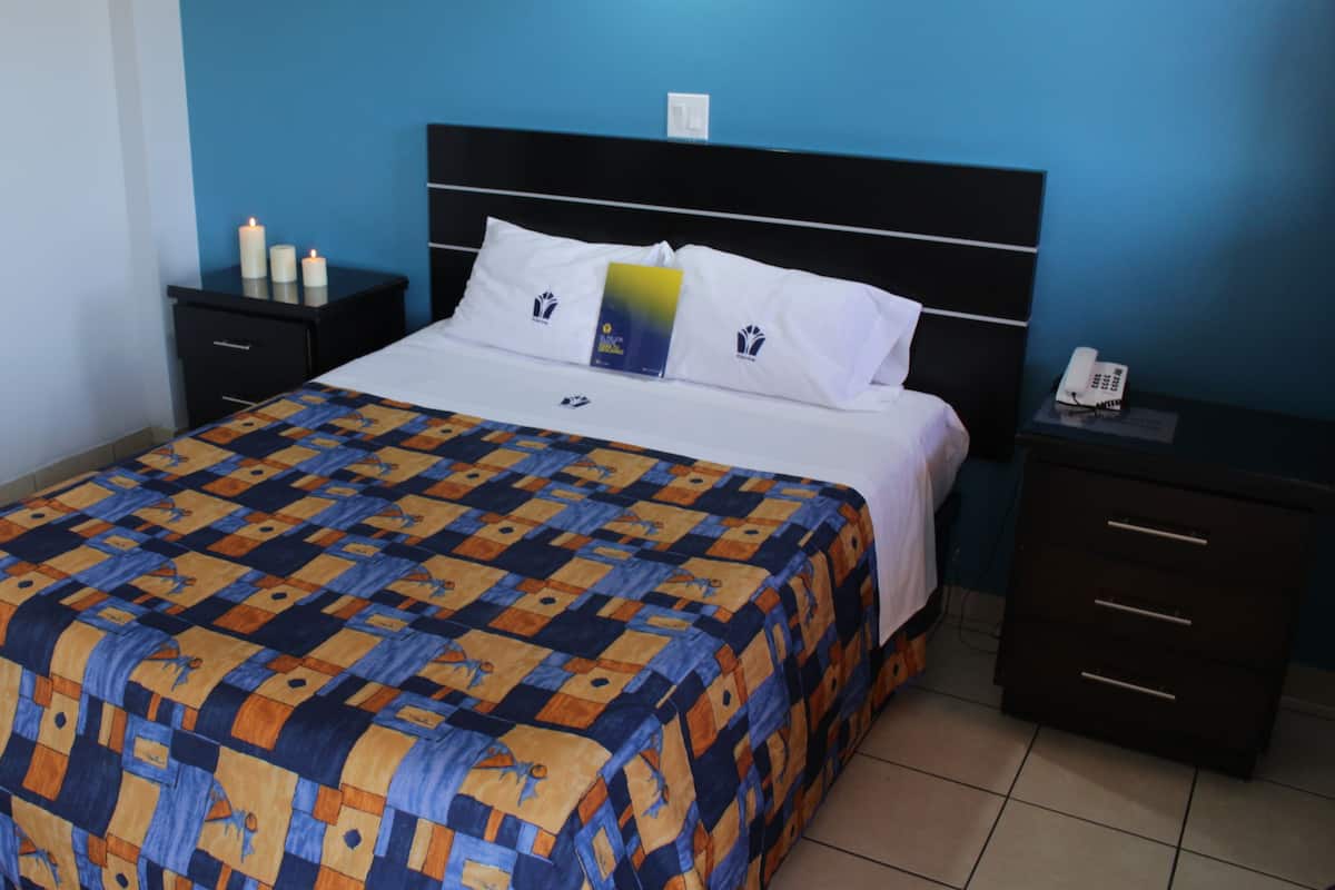 Una cama bien tendida con una colcha estampada, un cabecero negro y una mesita de noche con un teléfono y un libro.