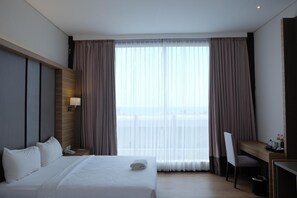 Quarto de hotel com cama, escrivaninha, cadeira, abajur e vista através de cortinas translúcidas.