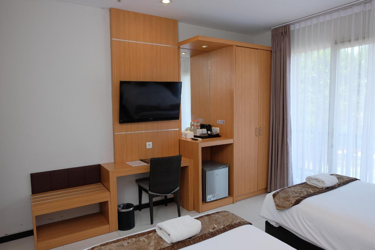 Quarto de hotel com escrivaninha de madeira, televisão, mini-geladeira, cama e janela com cortinas.