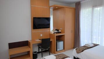 Quarto de hotel com escrivaninha de madeira, televisão, mini-geladeira, cama e janela com cortinas.