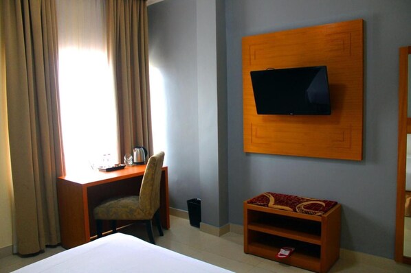 A hotel room with a bed, a desk with a chair, a television mounted on the wall, and a small wooden side table.