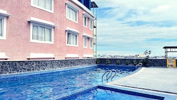 A swimming pool with a stone border, a pink building with white windows, and a clear sky.
