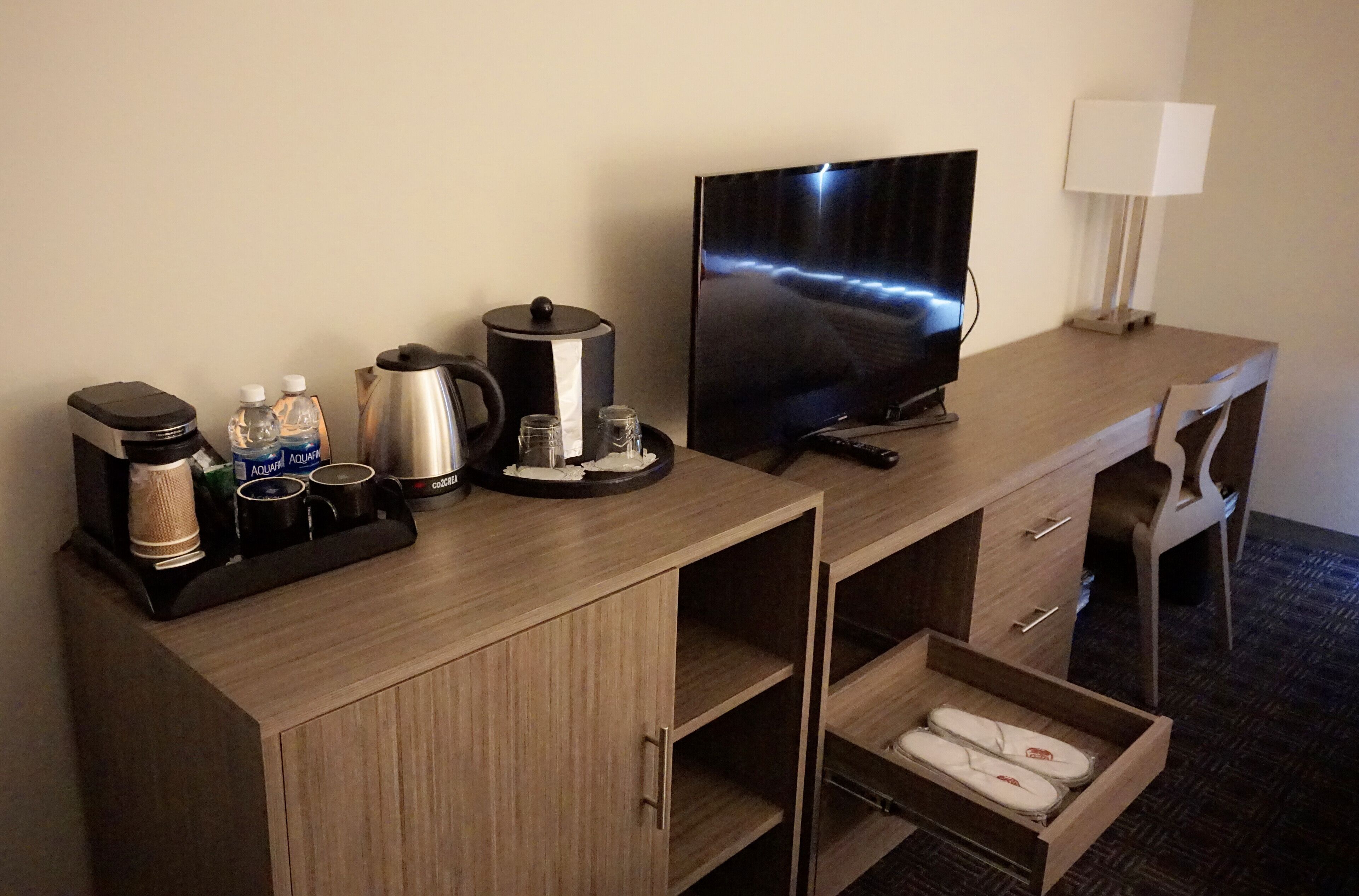 A hotel room desk with a television, coffee maker, water bottles, and a lamp.