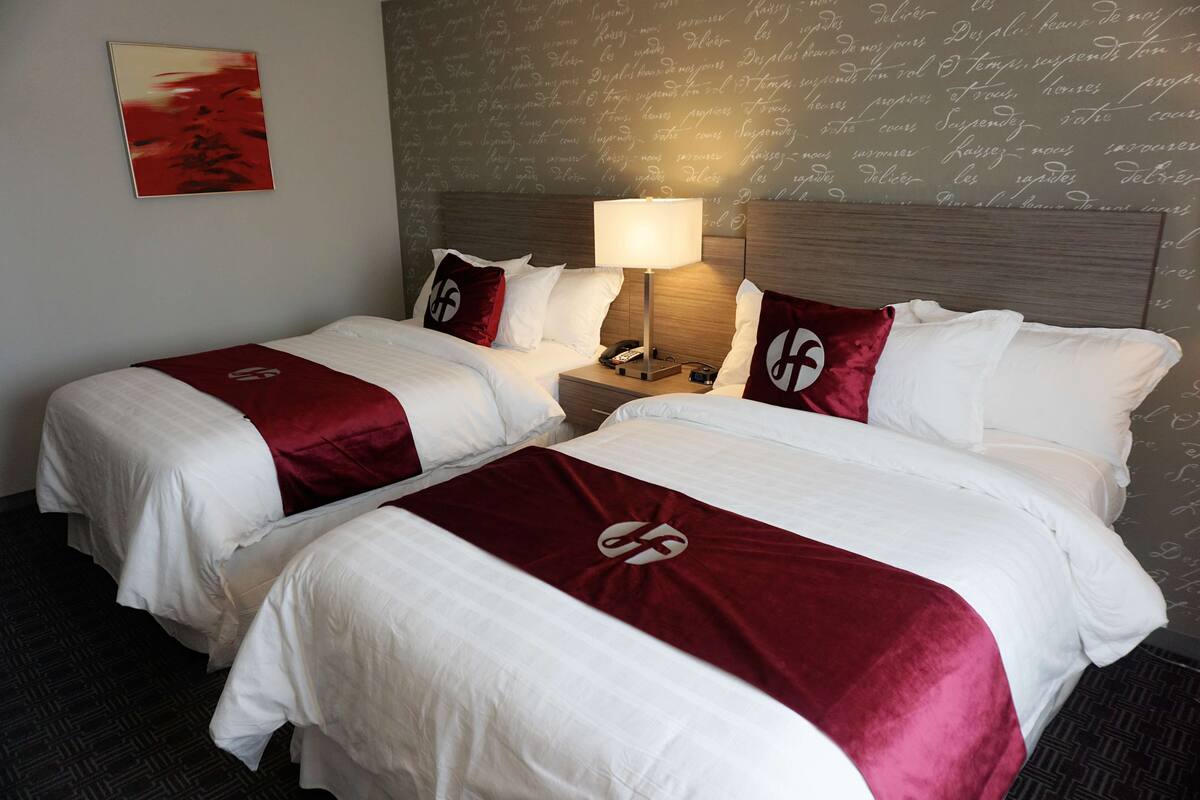 A hotel room with two beds, each with white bedding and red throw pillows, a bedside table with a lamp, and a wall with decorative script writing.