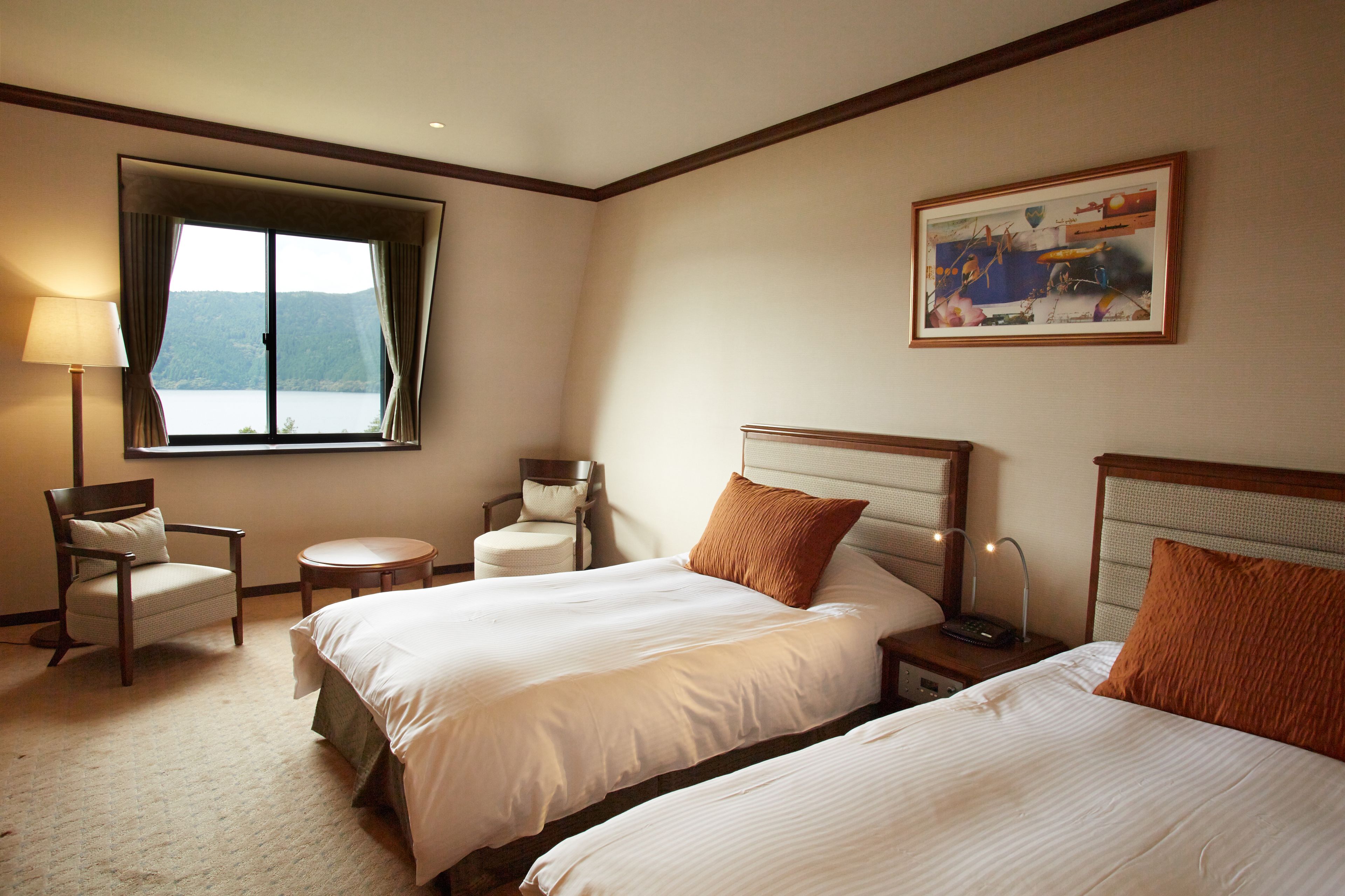 A hotel room with two beds, a chair, a small table, a lamp, and a painting on the wall.