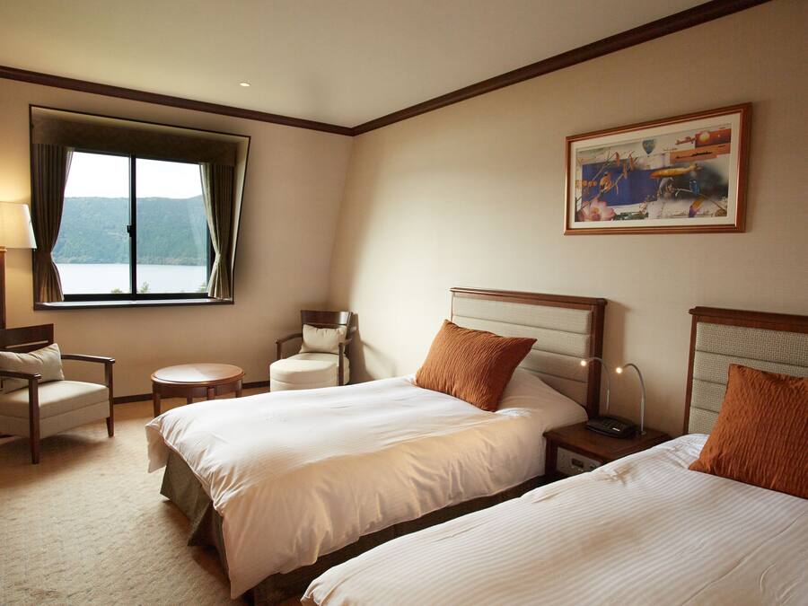 A hotel room with two beds, a chair, a small table, a lamp, and a painting on the wall.