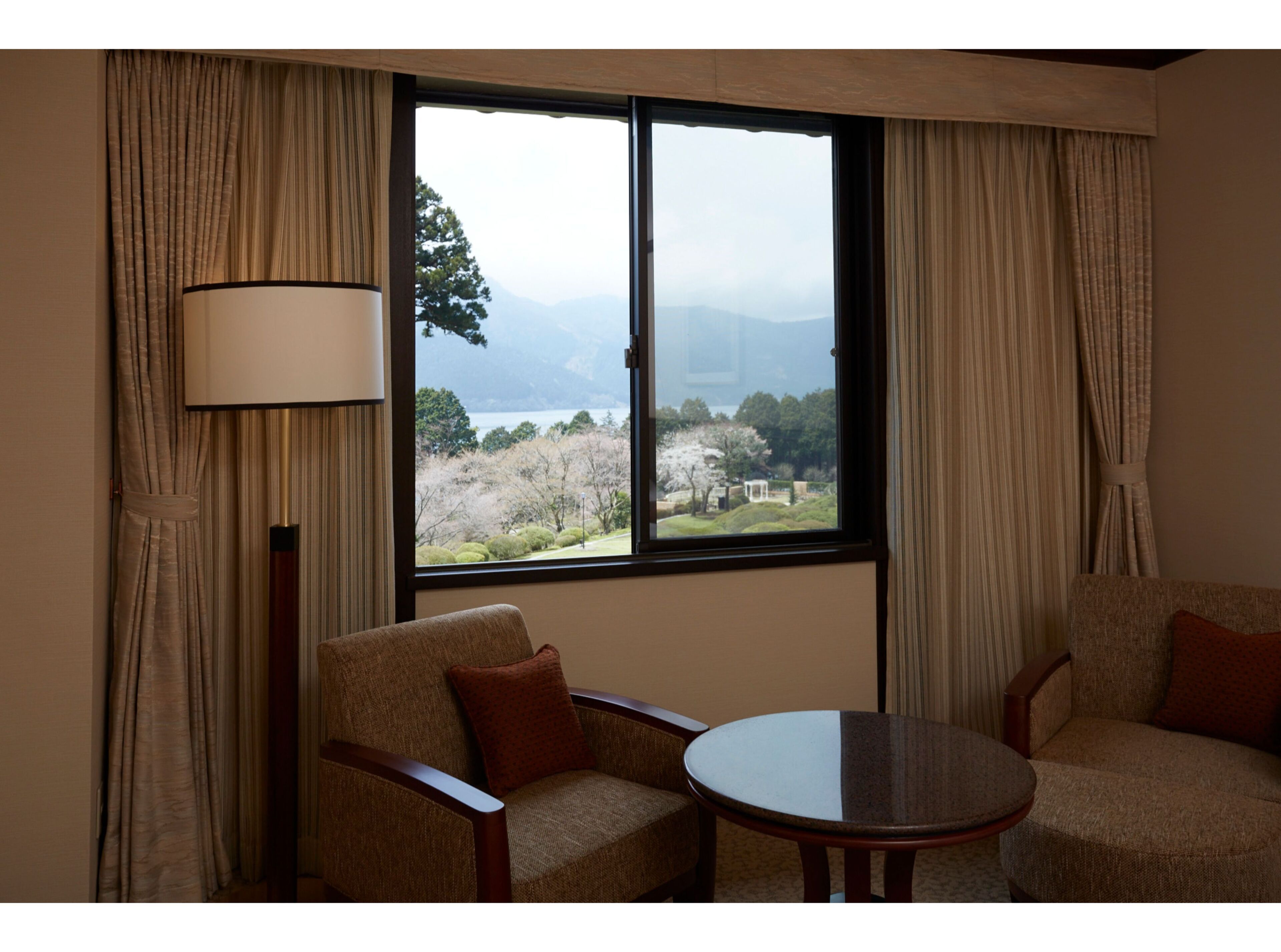 A room with a large window, two armchairs, a small round table, and a floor lamp.