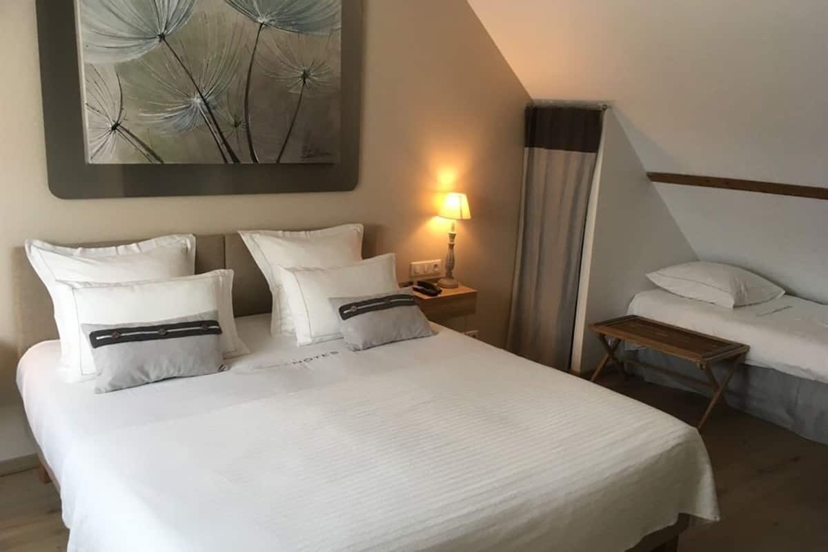 Habitación de hotel con una cama grande, una cama más pequeña, una mesita de noche con lámpara y un cuadro en la pared.
