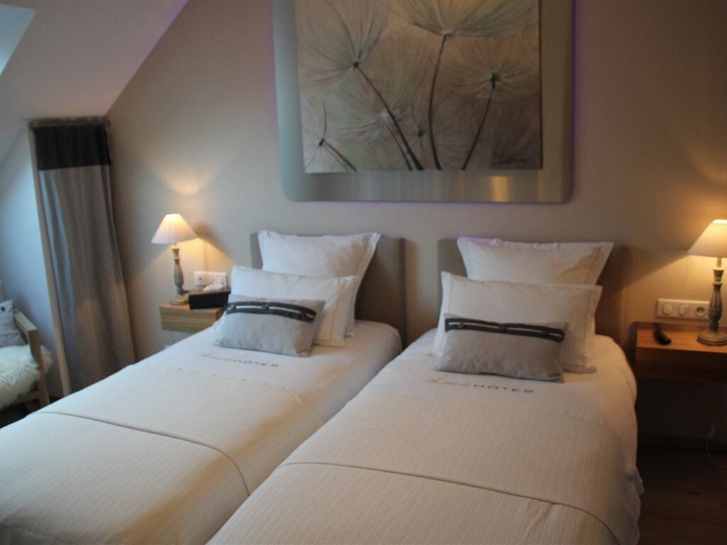 A hotel room with two beds, each with white bedding and a black stripe. There are bedside tables with lamps, a painting on the wall, and a window with curtains.