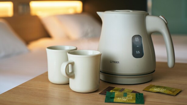 A bed with pillows and a bedside table with a kettle, two mugs, and packets of creamery powder.