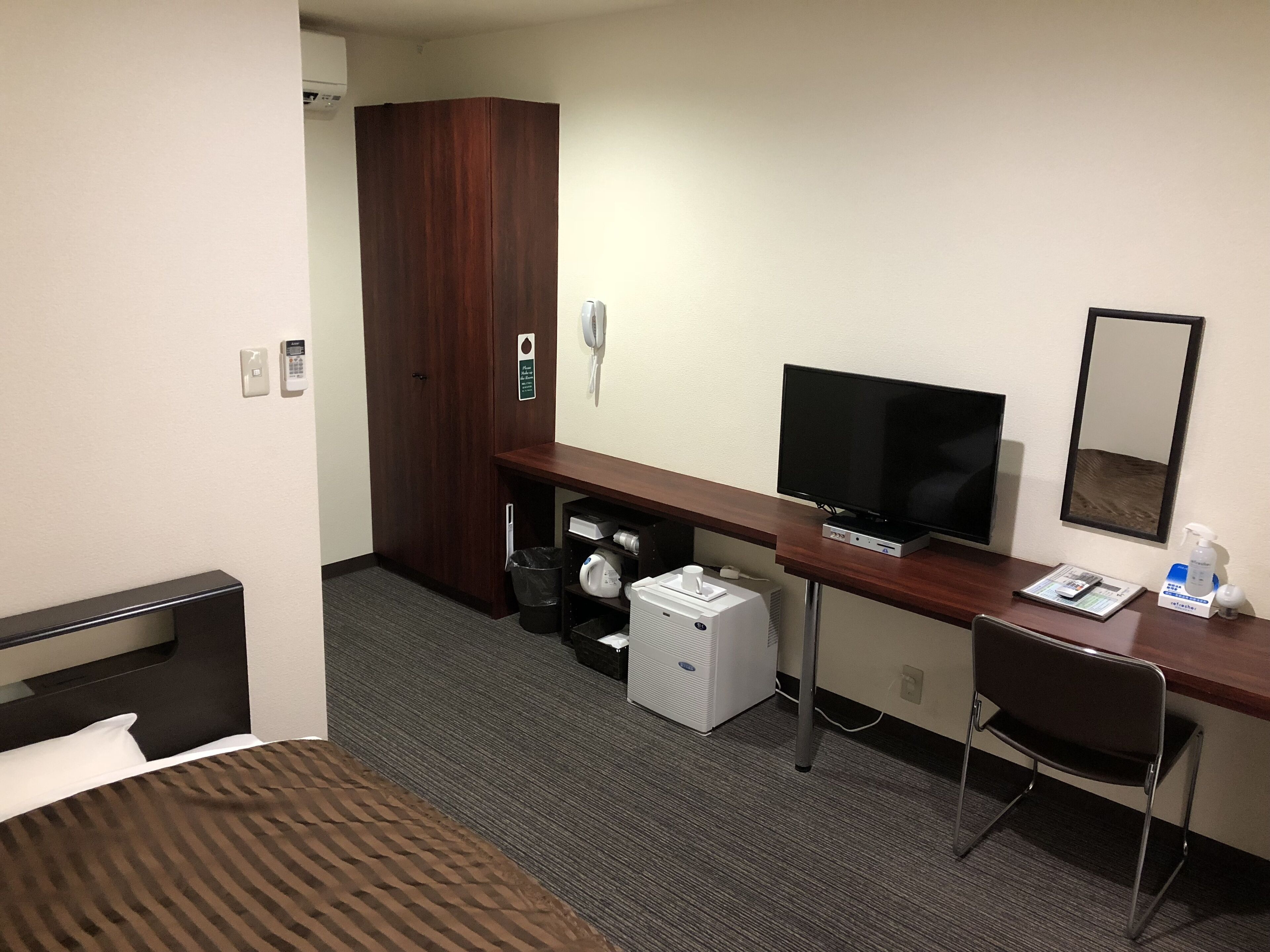 A hotel room with a bed, a desk with a TV, a chair, a wardrobe, a trash bin, and a small heater.