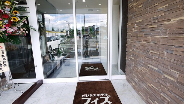 A reception area with a glass door, a brown mat with Japanese text, and a wooden wall. - Business Hotel Fiz Nagoya Airport (Toyoyama)