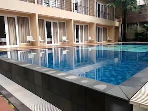 Una piscina con agua azul cristalina, rodeada por una solana empedrada y un edificio con balcones y ventanas.