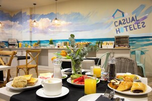 Una escena de desayuno con un mural costero, sillas de mimbre y una variedad de alimentos para el desayuno.