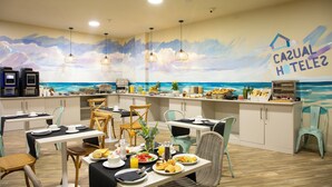 Un espace dédié au petit-déjeuner, agrémenté d’une fresque représentant une plage et l’océan, de tables où sont disposés des aliments pour le petit-déjeuner, et de luminaires suspendus.