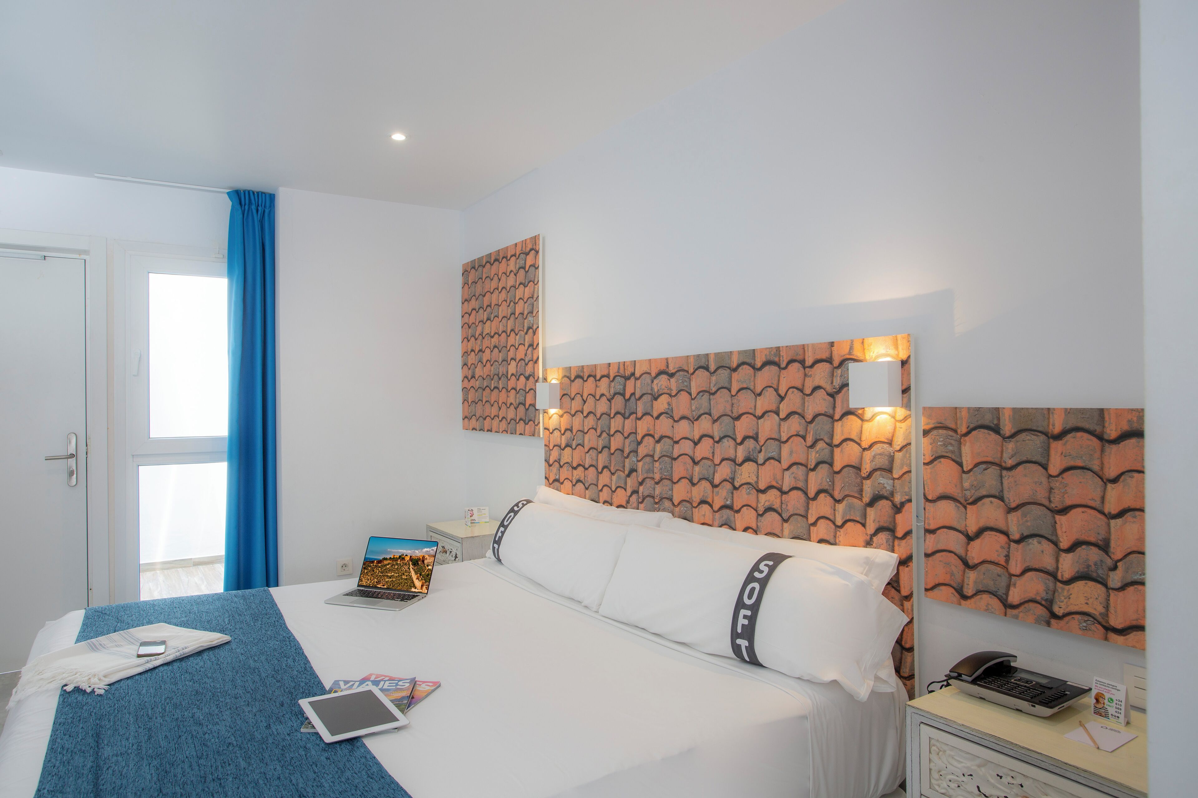 Habitación de hotel con una cama, una mesita de noche con lámpara, una computadora portátil, una tableta y un teléfono sobre la cama.