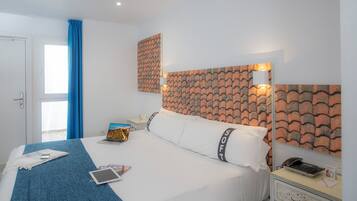 Habitación de hotel con una cama, una mesita de noche con lámpara, una computadora portátil, una tableta y un teléfono sobre la cama.