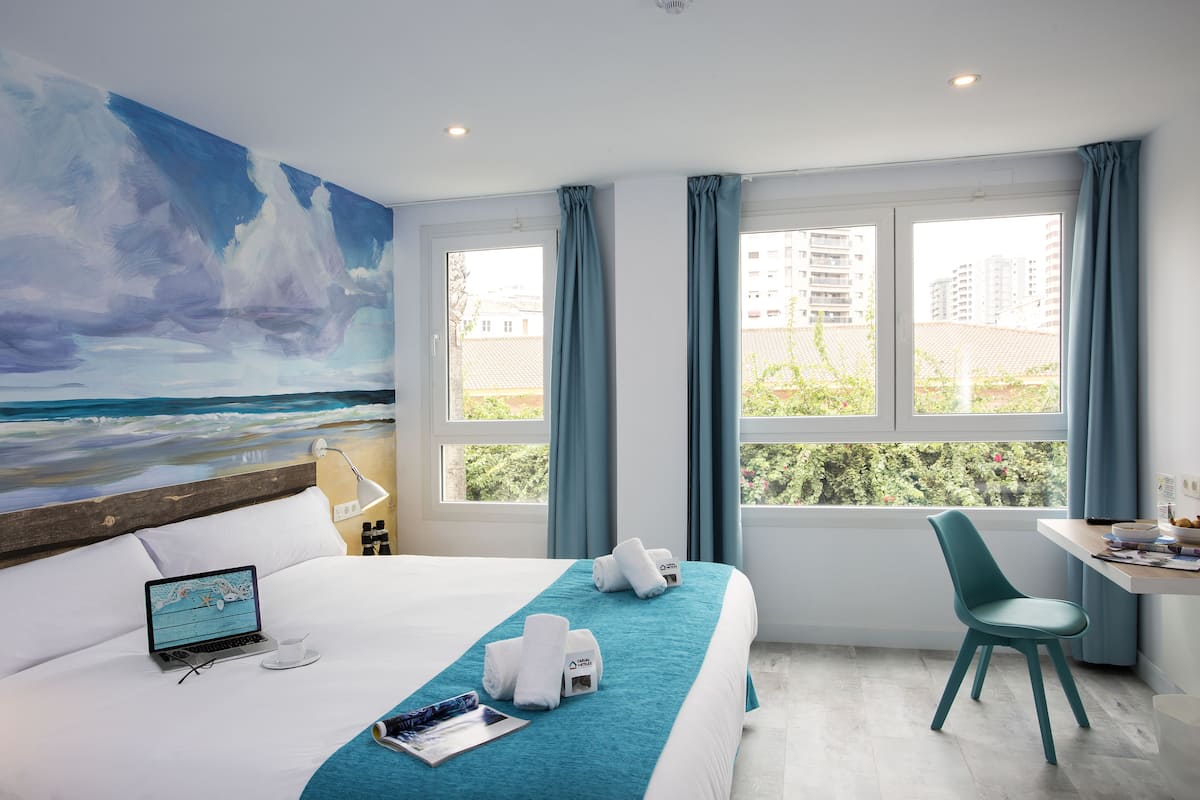 Een hotelkamer met een bed, een bureau, een stoel, een laptop en een schilderij van een strand.