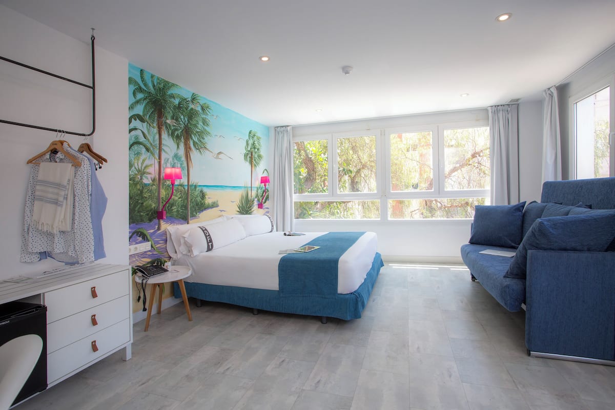 Een slaapkamer met een bed, een bureau, een stoel, een bank en een groot raam met uitzicht op palmbomen en de lucht.