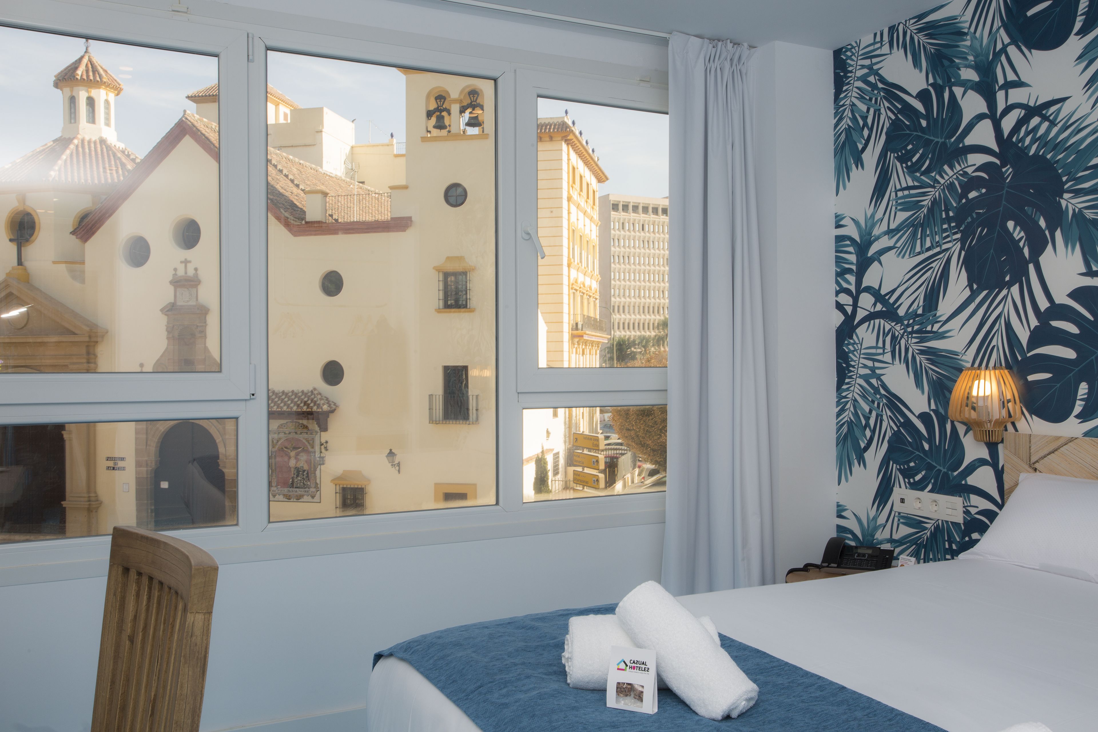 A hotel room with a bed, a chair, a window with a view of buildings, and a tropical wallpaper.