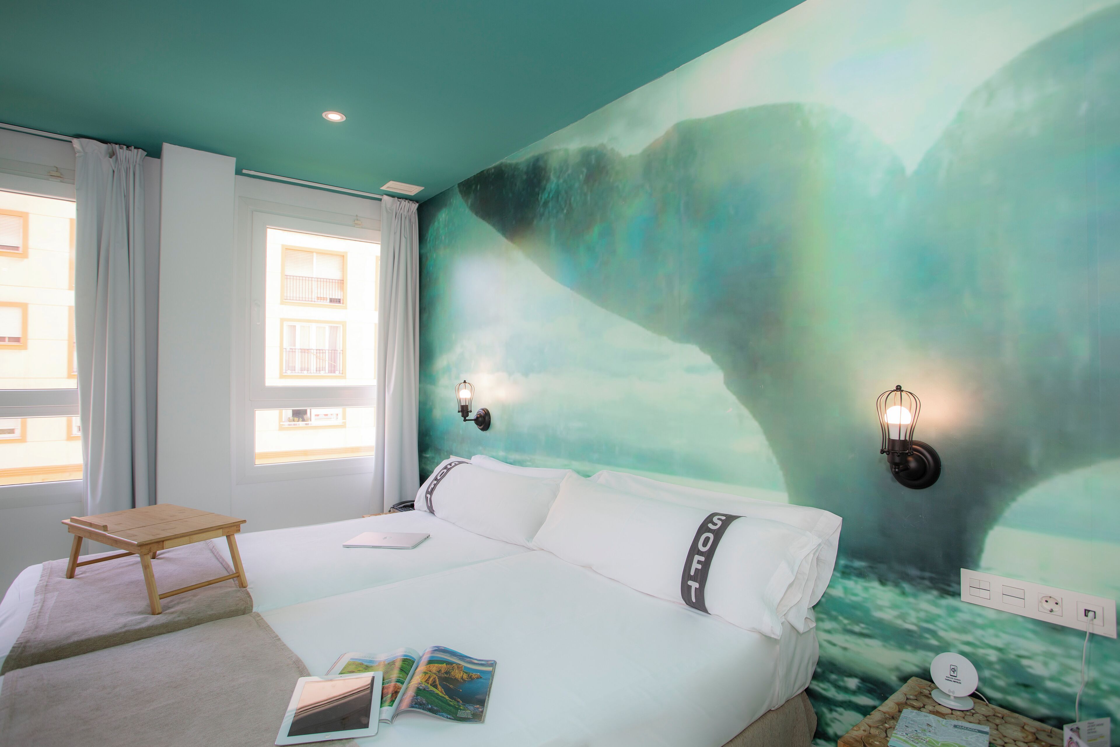 A hotel room with a bed, a small wooden table, a wall-mounted light, and a mural.