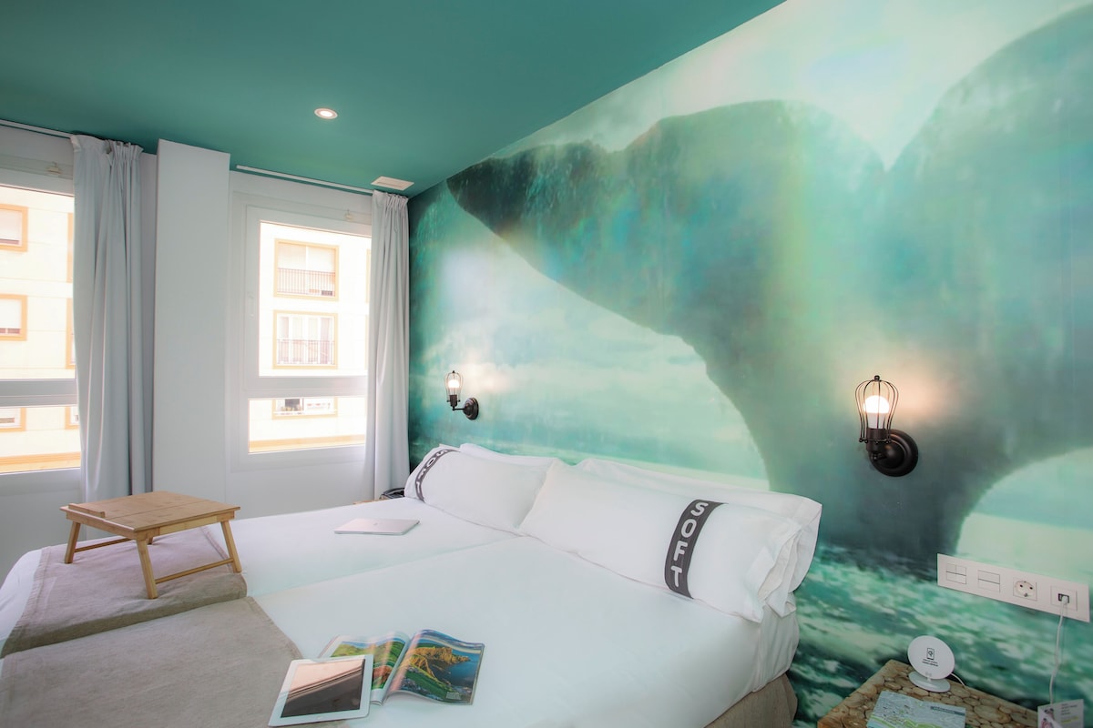 Een hotelkamer met een bed, een klein houten tafeltje, een wandlamp en een muurschildering.