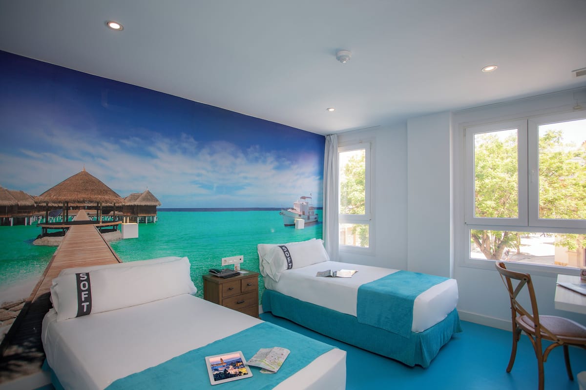 Een kamer met twee bedden, een bureau en een raam met uitzicht op een strand en waterbungalows.