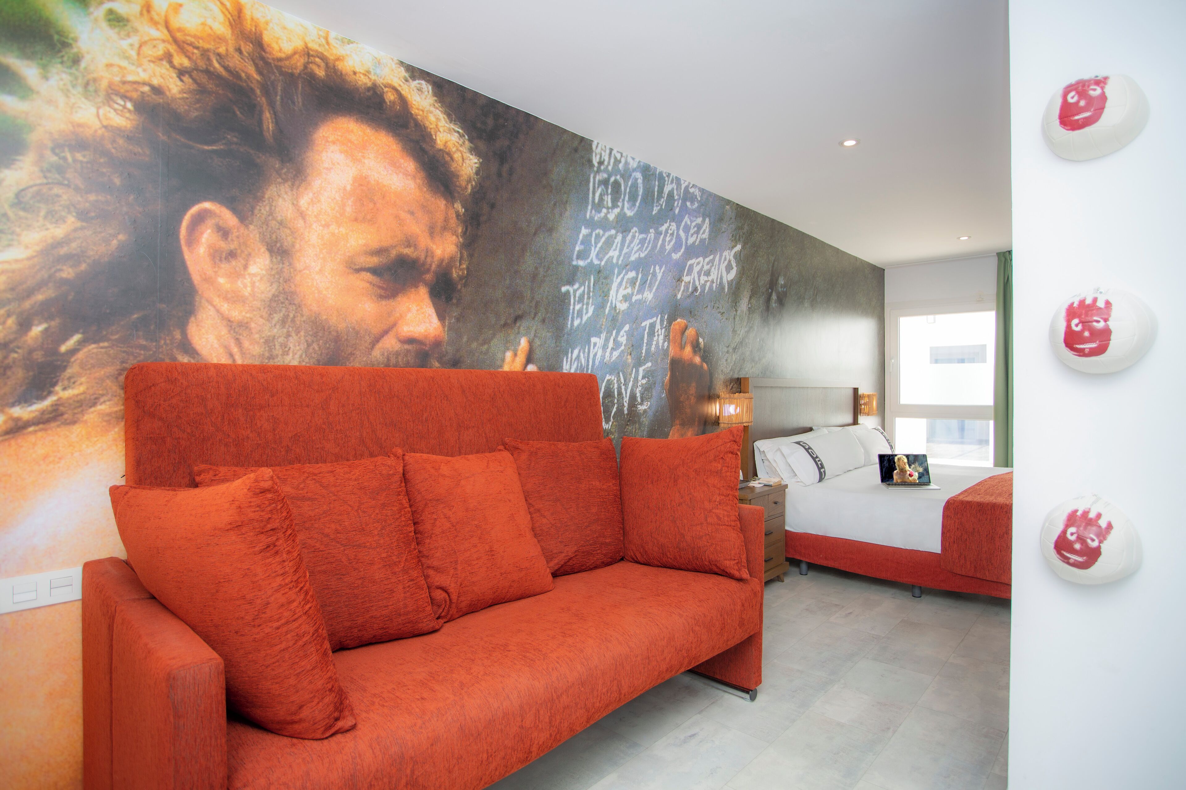 A modern bedroom with a red sofa, a bed, and a large mural on the wall.