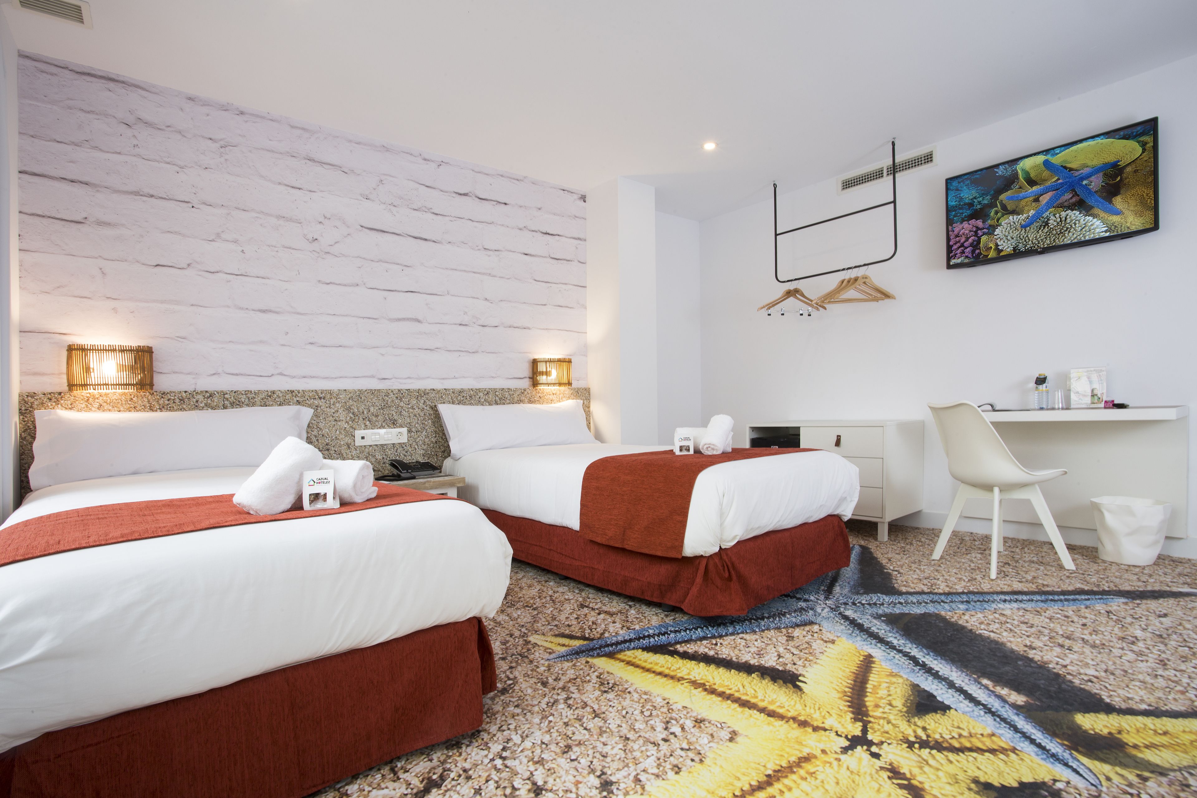 A hotel room with two beds, a desk, a chair, and a painting of an underwater scene.