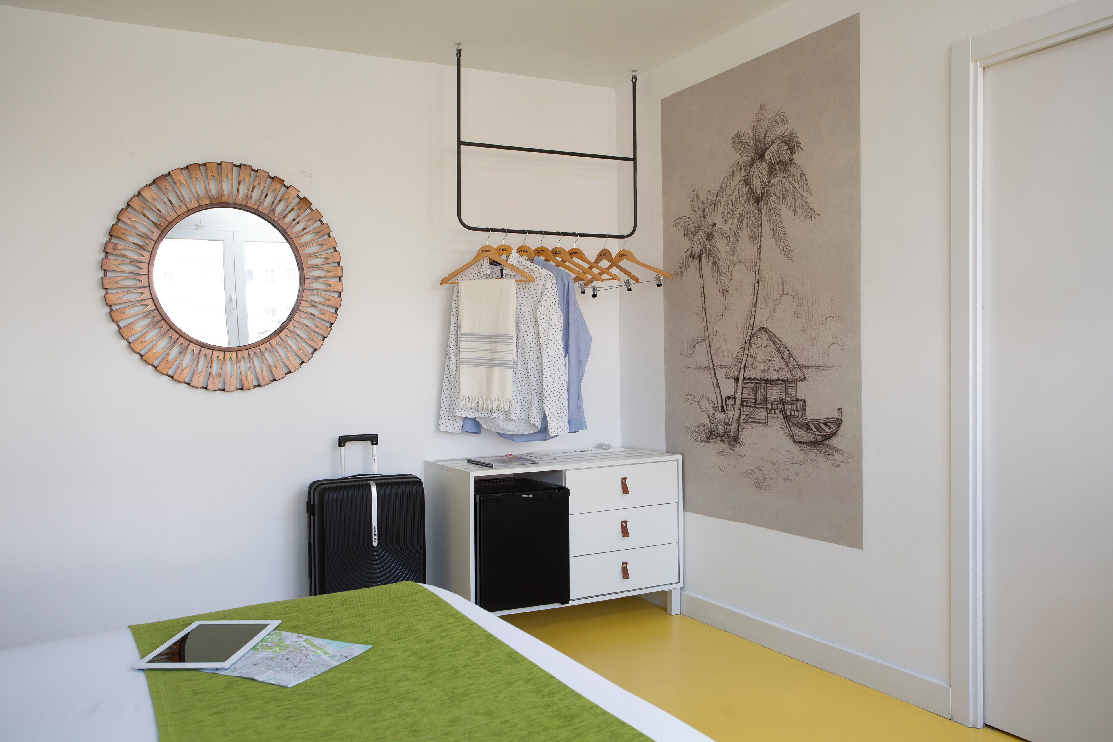 A hotel room with a bed, a desk, a small black refrigerator, a white dresser, a mirror, and a wall with a tropical mural.