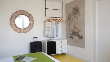 A hotel room with a bed, a desk, a small black refrigerator, a white dresser, a mirror, and a wall with a tropical mural.