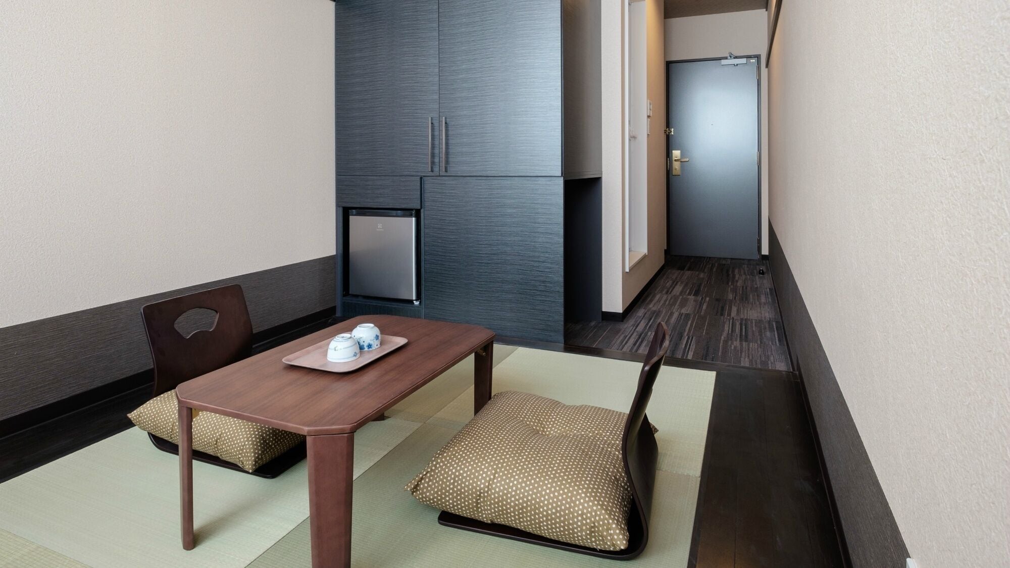 A small hotel room with a wooden table, two chairs, a refrigerator, and a door.