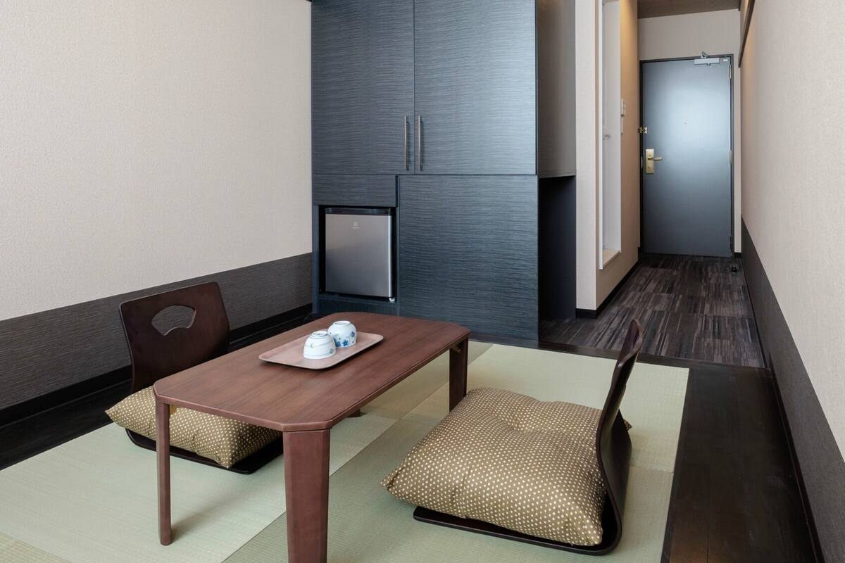 A small hotel room with a wooden table, two chairs, a refrigerator, and a door.
