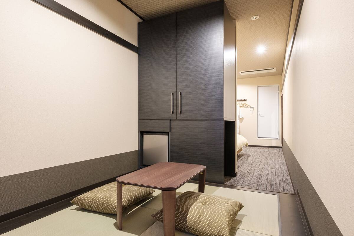 A compact hotel room with a small table, two pillows, a wardrobe, and a bed.